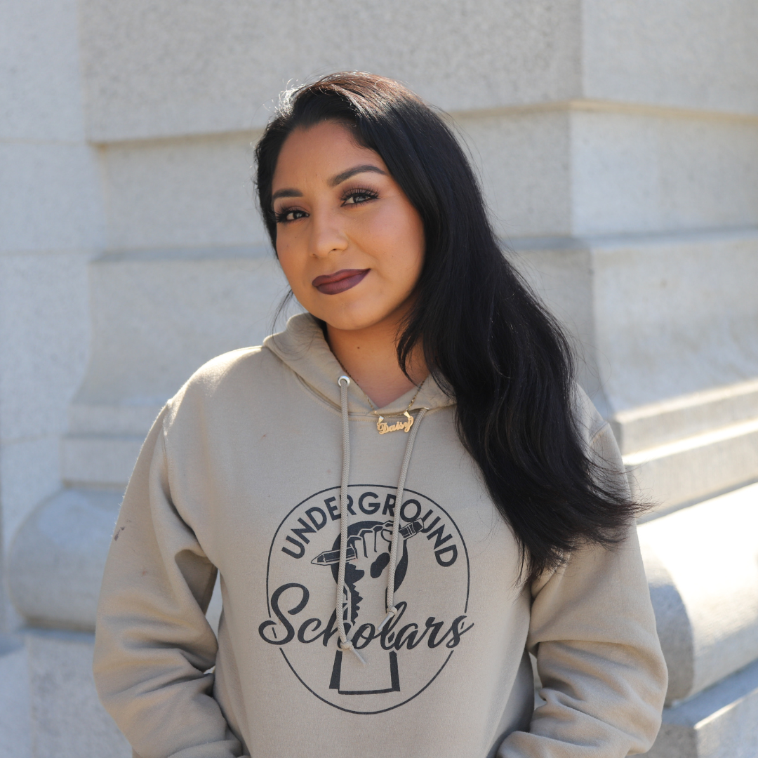 2023 Democracy Camp in DC Fellow Daisy Flores; young woman with wavy, brown hair past her shoulders standing outside in a tan hoodie with black letters saying "Underground Scholars"