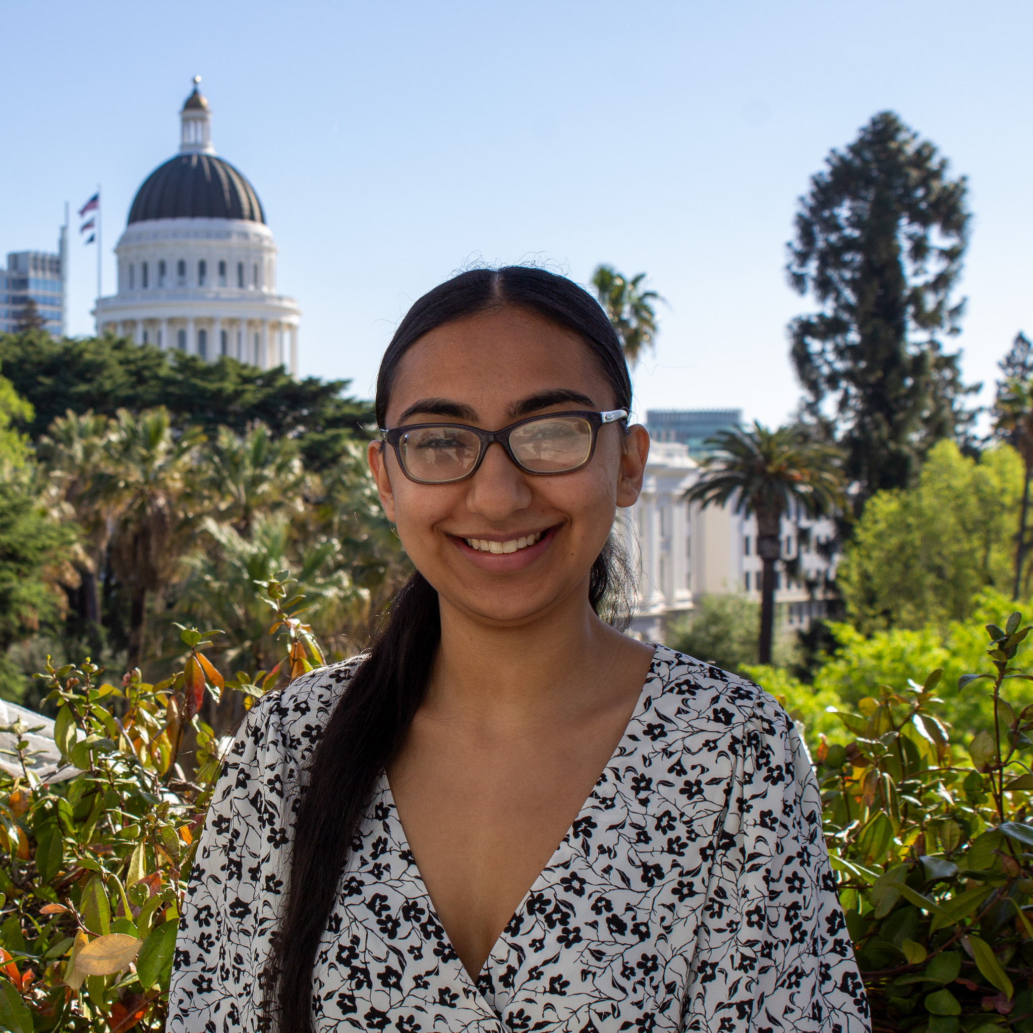 young woman with long brown hair in a ponytail and glasses standing in a white floral print blouse with the California State Capitol in the background
