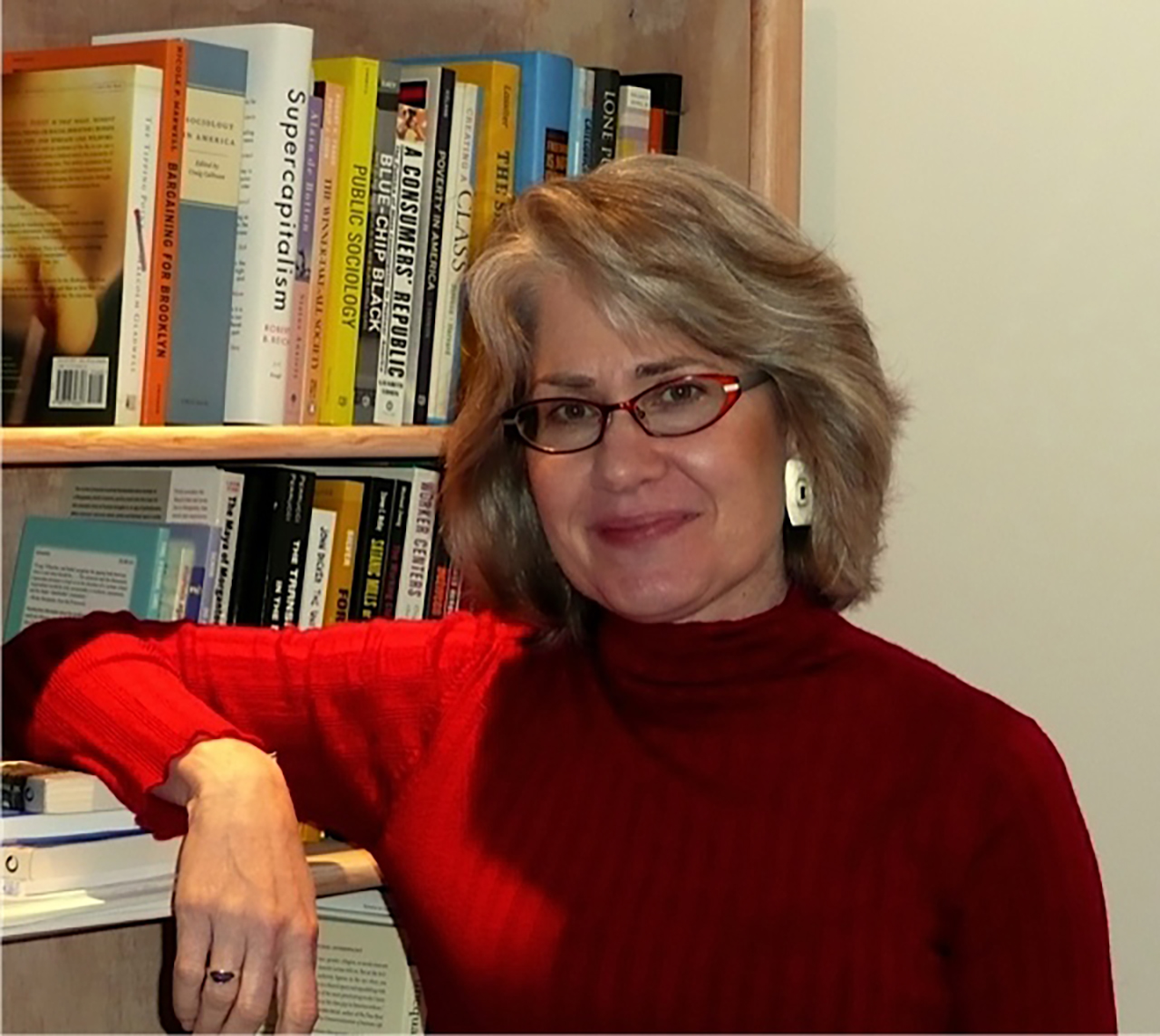 Kim Voss, posing in a red turtleneck leaning with her arm on a bookshelf