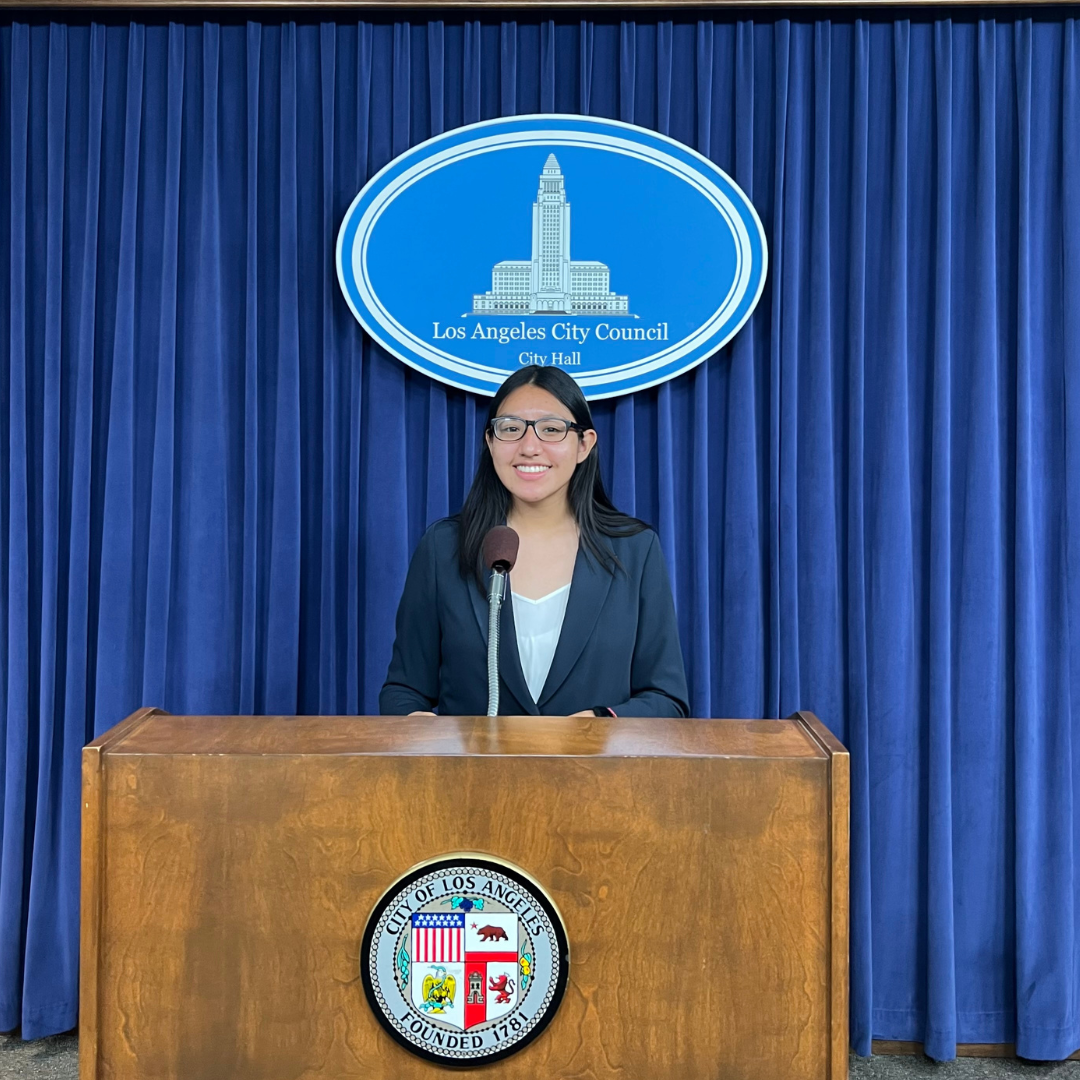 young woman in blazer in front of podium with the logo of Los Angeles City Council City Hall behind her