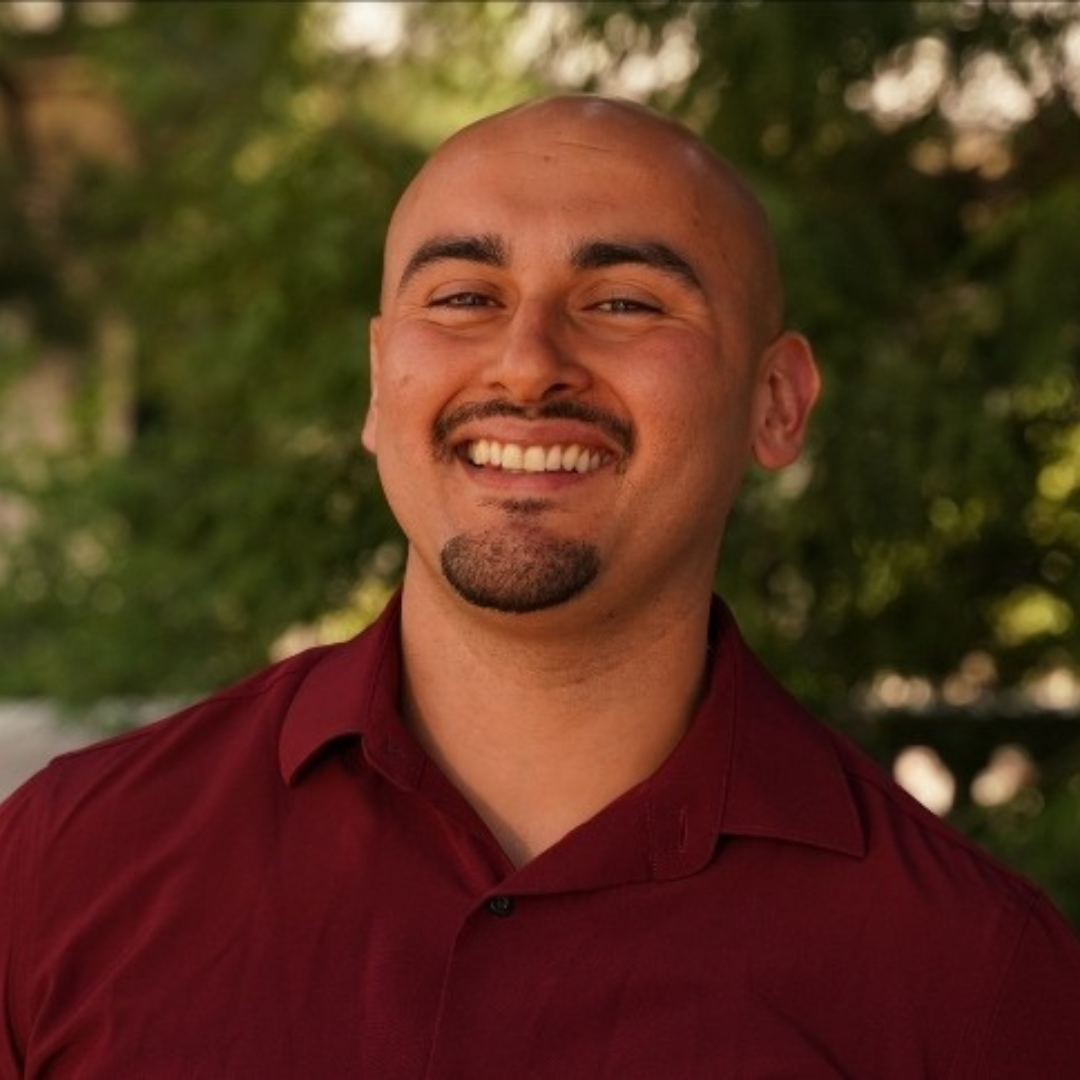 2023 Gardner Fellow Johnathan Lorenzo Dena bald man with a mustache and goatee stands smiling in a red buttondown shirt