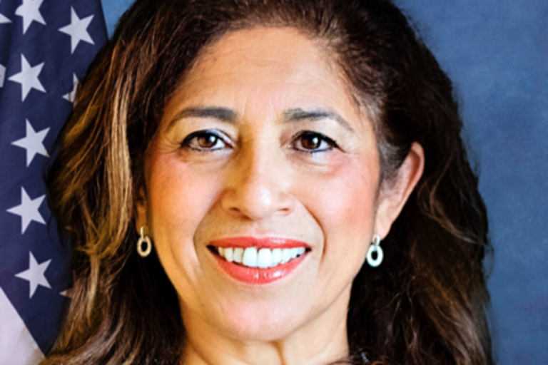 Elmy Bermejo SBA Administrator woman with shoulder length brown hair in front of blue background with American flag on the left