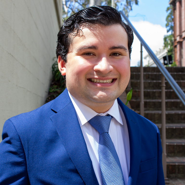 A young man in a blue suit smiles for a photo outside on a sunny day