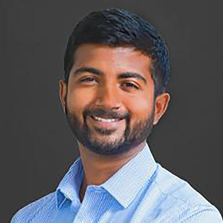 Deepak Premkumar smiling while wearing a blue collared buttondown against a dark gray background