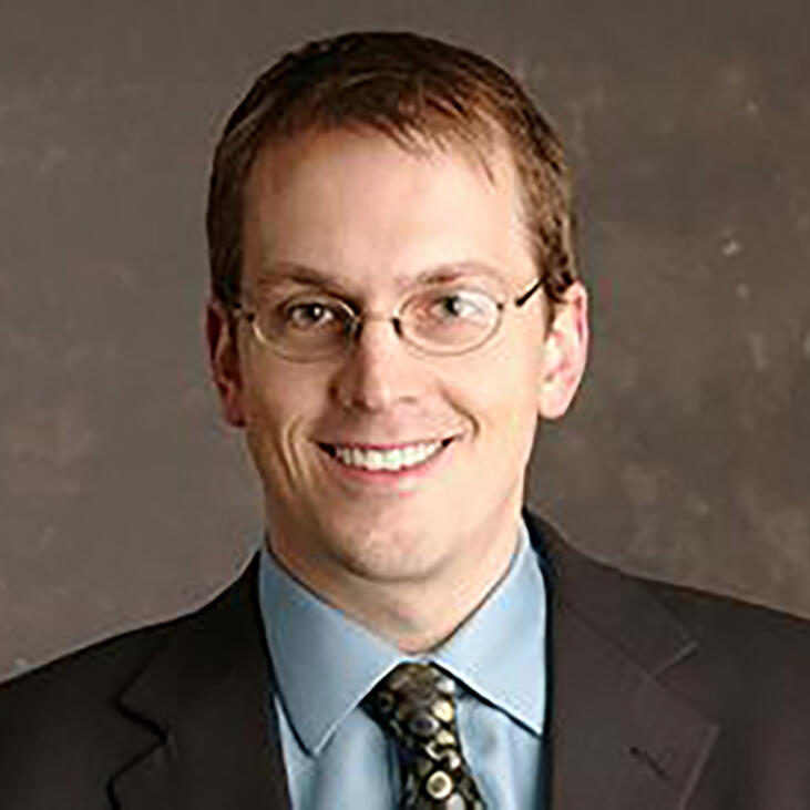 Eric McGhee, smiling in a dark suit, blue shirt, and gold patterned tie against a brown background