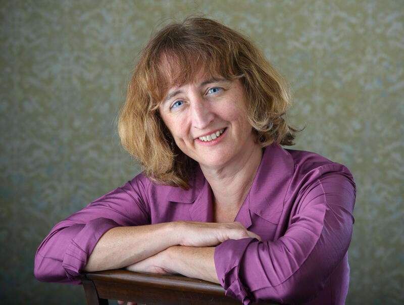 Karen Chapple in purble silk blouse, smiling with her arms folded and perched on the back of a chair