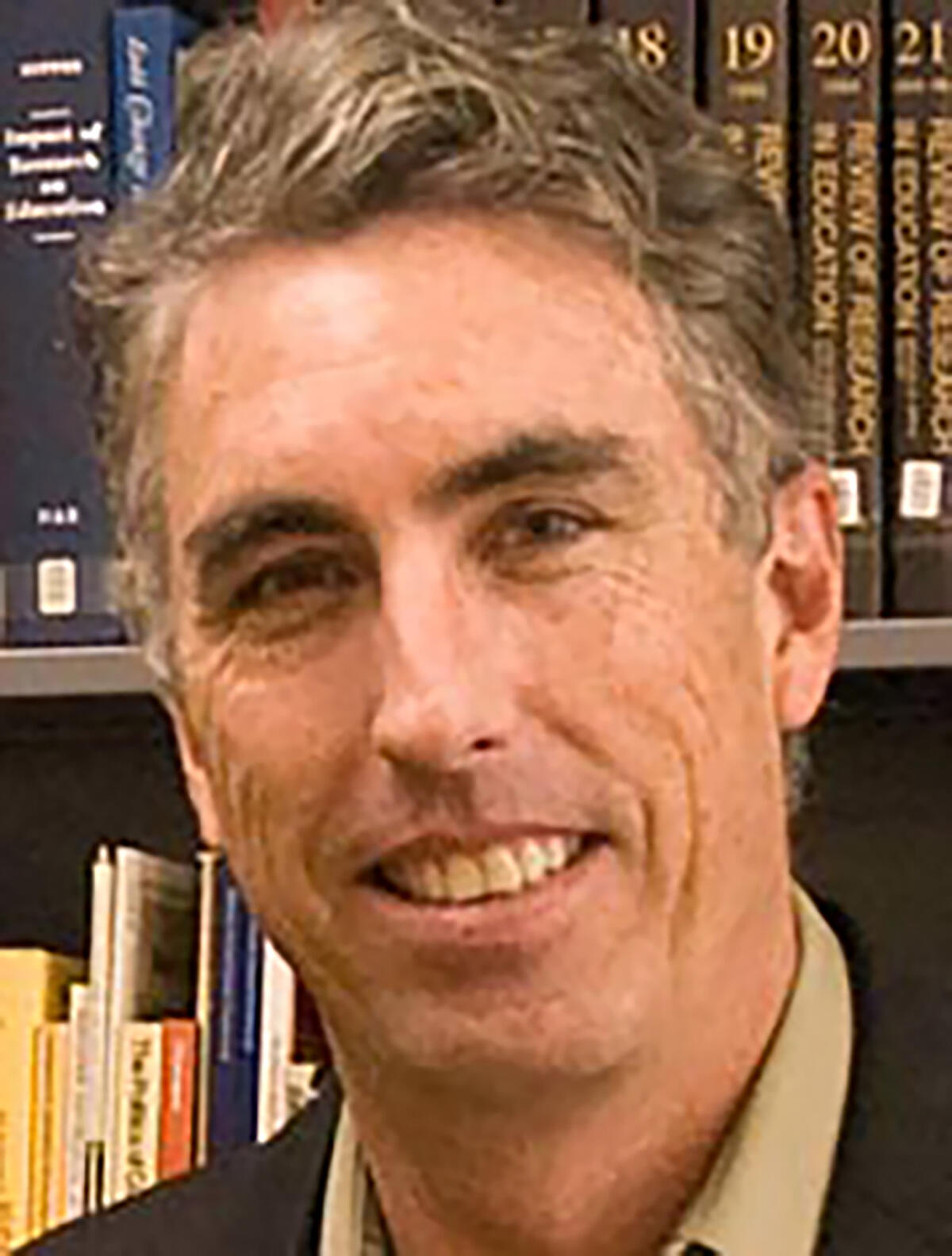 john aubrey douglass smiling in a beige buttondown and blue blazer in front of a bookshelf