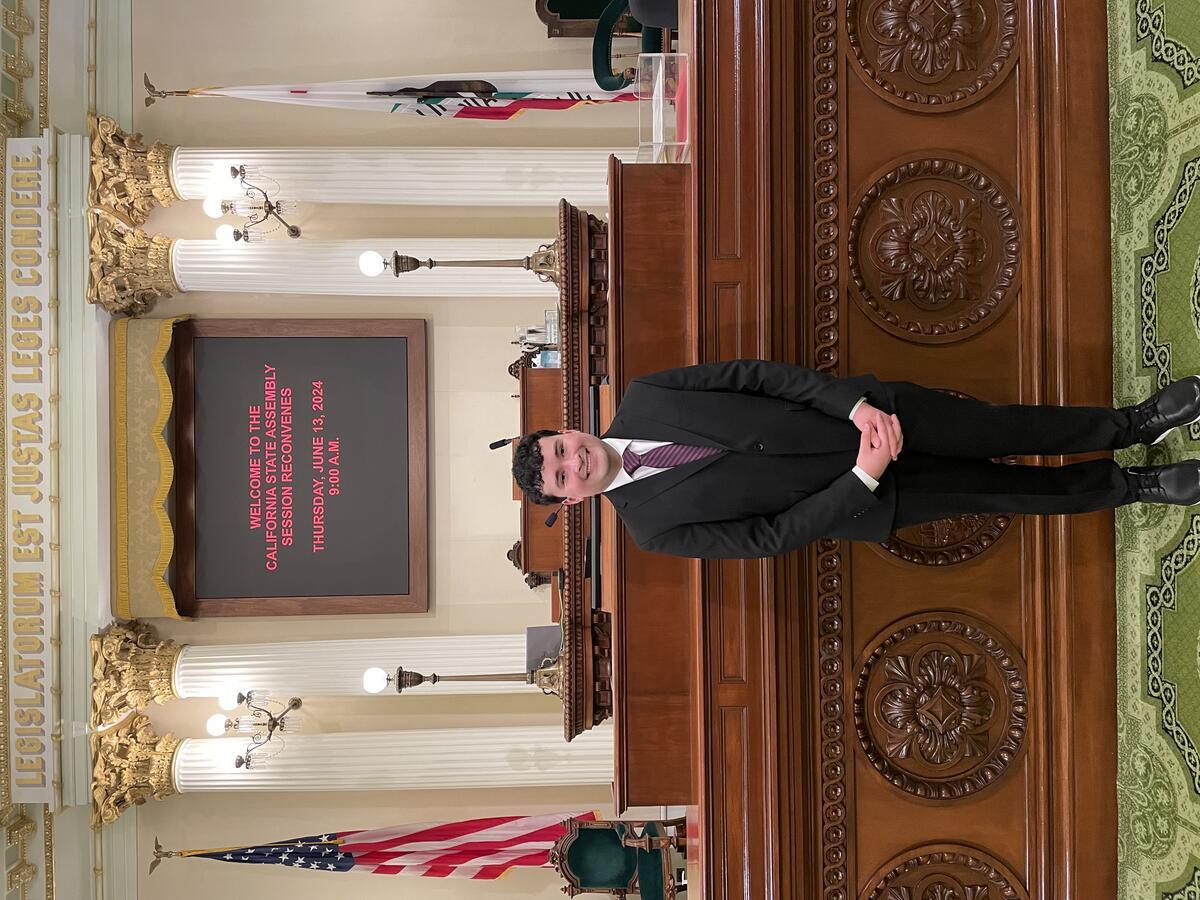 Man in professional clothing stands in front of an ornate podium with text in the background welcoming viewer to the California State Assembly;