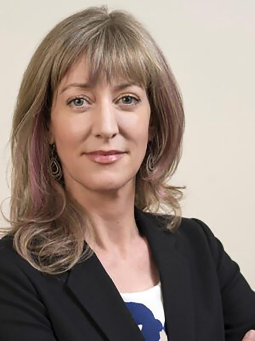 Ann Keller wearing a dark blazer and a white and blue patterned shirt against a beige background