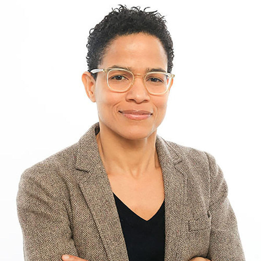 Nikki Jones smirking, wearing a tweed blazer and black v-neck shirt against a white background