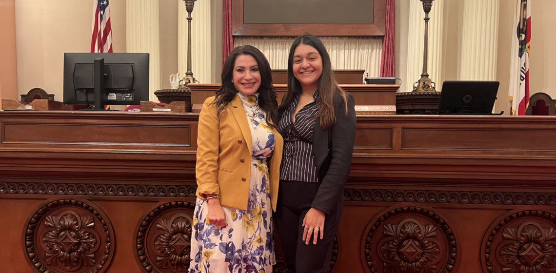 Two women in professional attire smile and pose for a photo in a formal interior space
