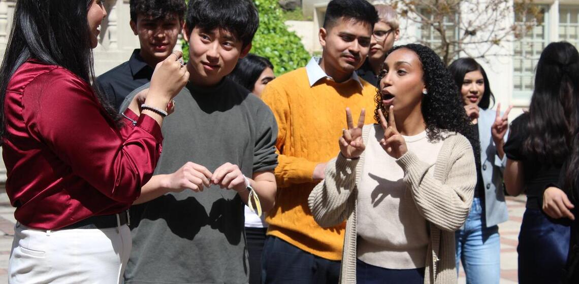 A group of young people stand outside and speak and gesticulate while holding up fingers