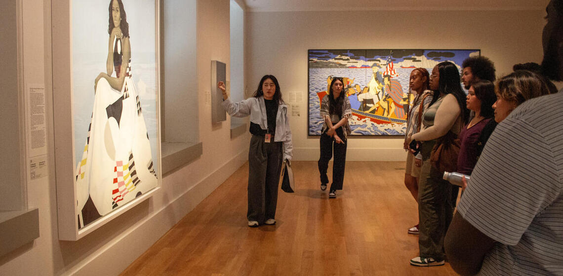 A group of people listen to a woman standing in front of a portrait of Michelle Obama