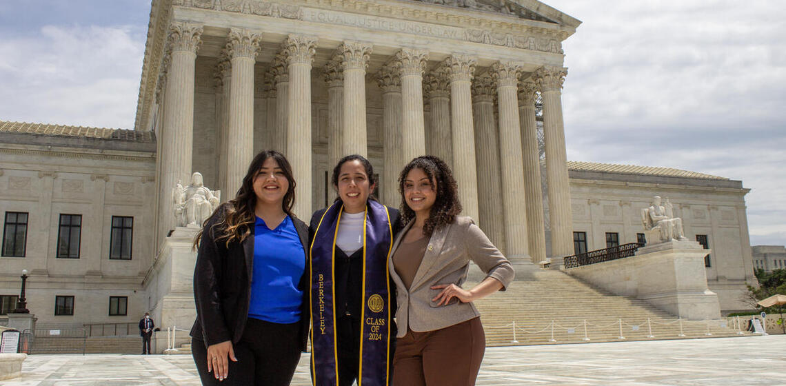 Three people in professional clothing in front of the US Supreme Court building. The center figure wears a graduation tassel.