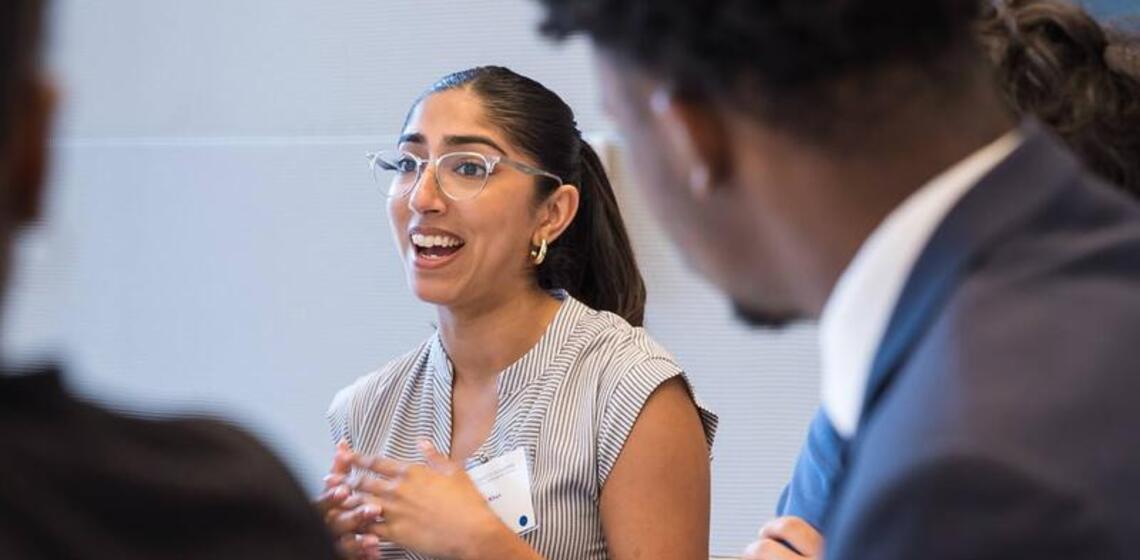 A young women in professional clothing speaking and gesticulating at a table in an interior space