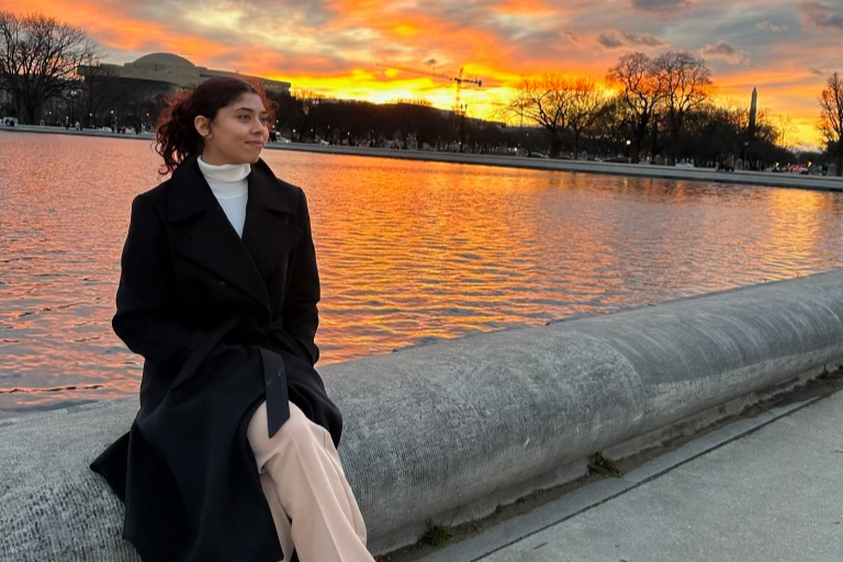 young woman sitting by the water in a black coat, beige pants, and white turtleneck